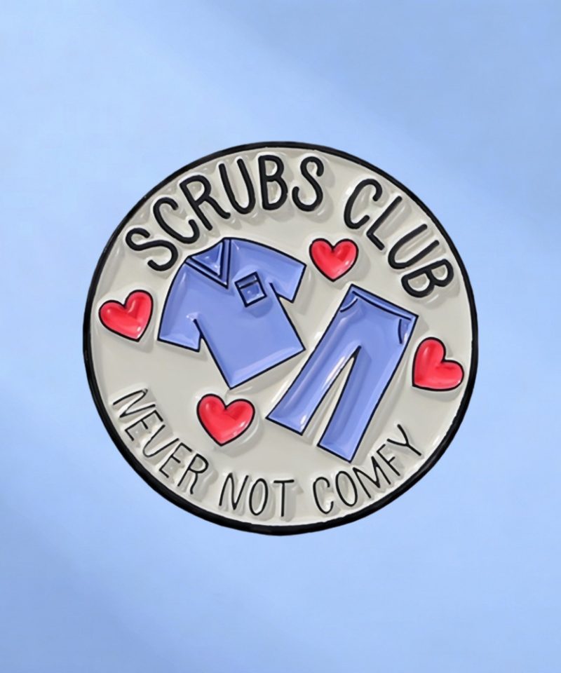 Round enamel pin reading Scrubs Club, Never Not Comfy, with a scrub set and red hearts