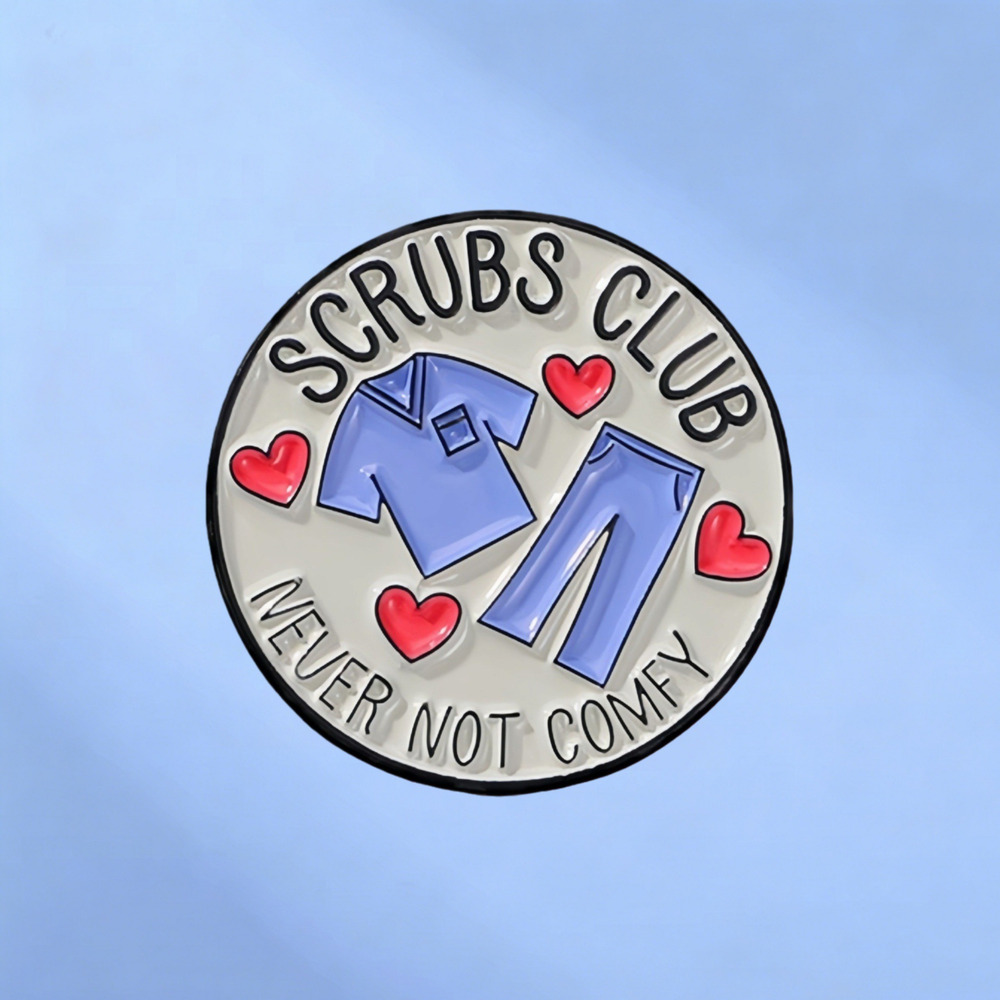 Round enamel pin reading Scrubs Club, Never Not Comfy, with a scrub set and red hearts