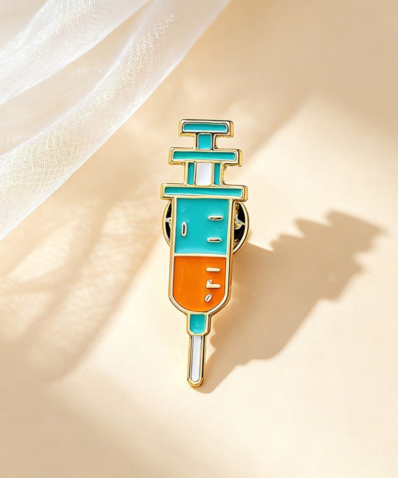 Teal and orange syringe enamel pin with a gold outline